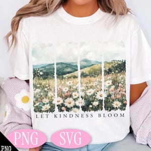 Op de afbeelding: Wit T-shirt met een aquarelontwerp van een bloemenveld en glooiende heuvels. De tekst "Let Kindness Bloom" is onder de afbeelding gedrukt.