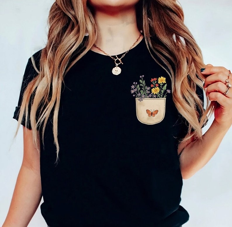 Puede incluir: Camiseta negra con un bolsillo con una mariposa y flores de colores.