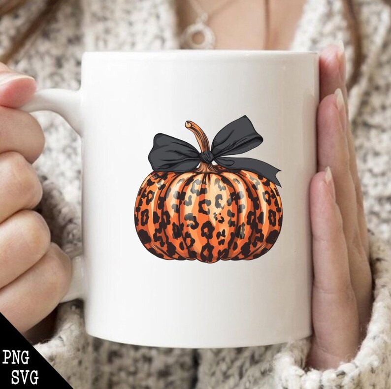 May include: White ceramic mug featuring a pumpkin design. The pumpkin is orange with black leopard spots and a black bow. The mug is held by a person wearing a white sweater.