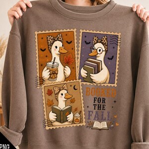 May include: A brown crewneck sweatshirt featuring a graphic print of three postage stamp-style illustrations of ducks. The ducks wear patterned headbands and are depicted holding books, a coffee cup, and a pumpkin mug. The text reads BOOKED FOR THE FALL.