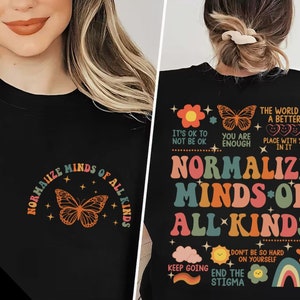 Puede incluir: Camiseta negra con un diseño de arcoíris que dice "Normalizar las mentes de todas las clases". El diseño incluye mariposas, estrellas, nubes y un arcoíris. El texto "Está bien no estar bien", "Eres suficiente", "El mundo es un lugar mejor contigo en él", "No seas tan duro contigo mismo", "Sigue adelante" y "Termina con el estigma" también están incluidos en el diseño.