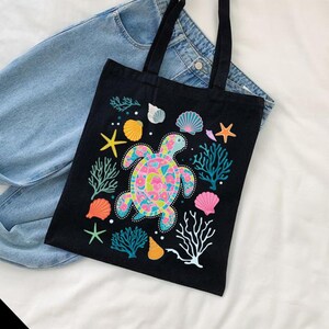 May include: Black canvas tote bag with a colorful sea turtle design. The turtle is surrounded by coral, seashells, and starfish.