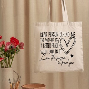 May include: A white canvas tote bag with black text that reads "Dear person behind me the world is a better place with you in it Love, the person in front you". The text includes a heart symbol.