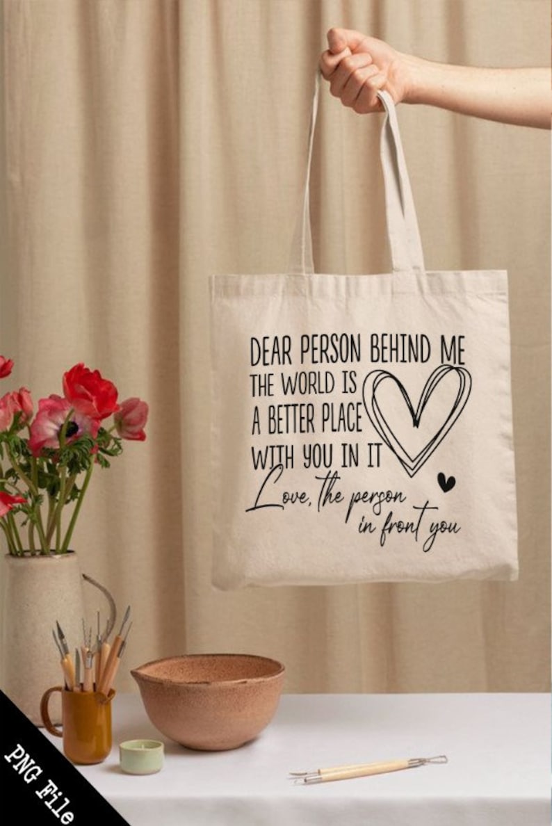 May include: A white canvas tote bag with black text that reads "Dear person behind me the world is a better place with you in it Love, the person in front you". The text includes a heart symbol.