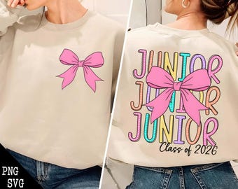 May include: A beige sweatshirt with a pink bow on the front and the text "Junior Junior Junior Class of 2026" on the back. The text is in rainbow colors with white polka dots.
