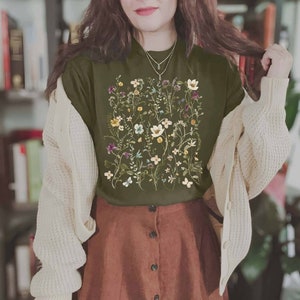 Puede incluir: Una camiseta verde con estampado floral que presenta una variedad de flores y hojas. La camiseta se combina con una falda marr&oacute;n y una chaqueta blanca de punto.