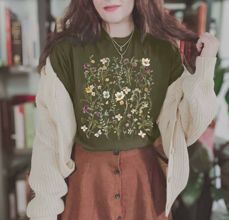 Puede incluir: Una camiseta verde con estampado floral que presenta una variedad de flores y hojas. La camiseta se combina con una falda marr&oacute;n y una chaqueta blanca de punto.