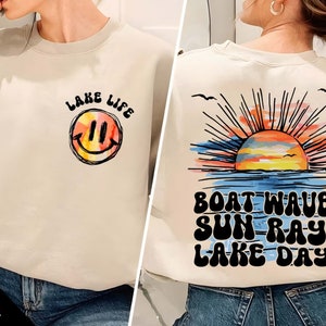May include: A light beige sweatshirt with a smiley face and the text "Lake Life" on the front. The back of the sweatshirt has a sunset graphic with the text "Boat Waves Sun Rays Lake Days".