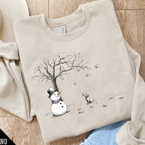 May include: A beige sweatshirt with a snowman and a dog illustration. The snowman is wearing a black hat and a scarf. The dog is looking at the snowman. The illustration is in black and white and is set against a background of a bare tree and falling leaves.