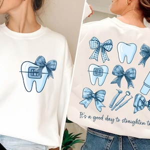 Puede incluir: Sudadera blanca con un diente azul con brackets y un lazo azul en la parte delantera. La parte posterior de la sudadera tiene un lazo azul, un diente con brackets, un lazo azul, un diente, un lazo azul, un diente, un lazo azul, un cepillo de dientes azul, una taza azul y un lazo azul. El texto "Es un buen día para enderezar los dientes" está impreso en la parte posterior de la sudadera.