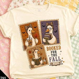 May include: A cream-colored t-shirt featuring a graphic of three postage stamp-style illustrations of ducks. The ducks wear patterned bows and are depicted holding books or a beverage. The text reads BOOKED FOR THE FALL with autumn leaves.
