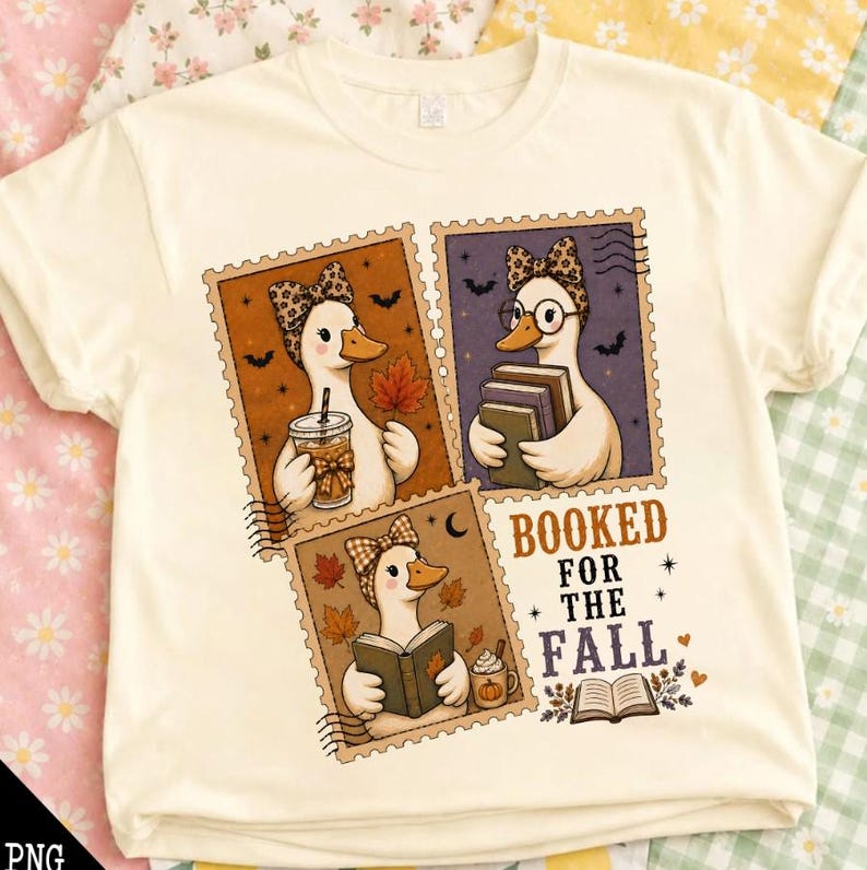 May include: A cream-colored t-shirt featuring a graphic of three postage stamp-style illustrations of ducks. The ducks wear patterned bows and are depicted holding books or a beverage. The text reads BOOKED FOR THE FALL with autumn leaves.