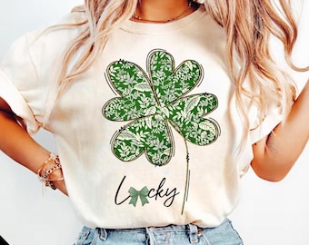 May include: A white t-shirt with a green four-leaf clover design and the word "Lucky" written in black with a green bow.