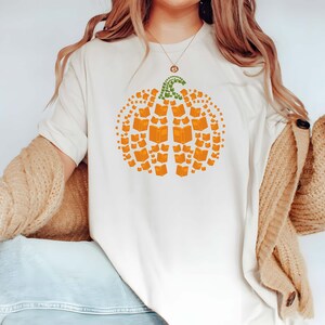 May include: A white t-shirt with a graphic of a pumpkin made of books. The pumpkin is orange and the stem is green.