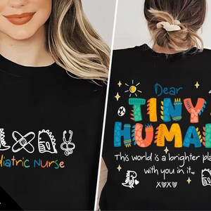May include: Two black t-shirts with colourful designs. One features the text "Pediatric Nurse" alongside medical symbols and dinosaur drawings. The other says "Dear TINY HUMAN" with a positive message and dinosaur illustrations.