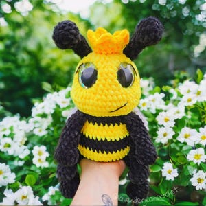 May include: A handmade crochet bee plushie with a yellow body, black stripes, and large, sparkly eyes. The bee has black antennae and a yellow crown. The plushie is held in front of a background of white flowers and green foliage.