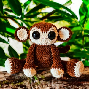 May include: A handcrafted crochet monkey toy in brown and white, featuring large black eyes and a smiling face. The soft toy has a textured, fluffy look and is positioned on a wooden surface. The monkey's ears, face, hands, and feet are white.