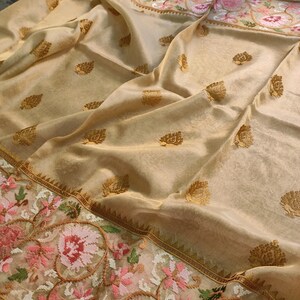 Pode incluir: Um sari bege com padr&otilde;es de folhas douradas e uma borda floral bordada em rosa, verde e branco. O tecido tem uma qualidade brilhante e transl&uacute;cida, com uma queda delicada.