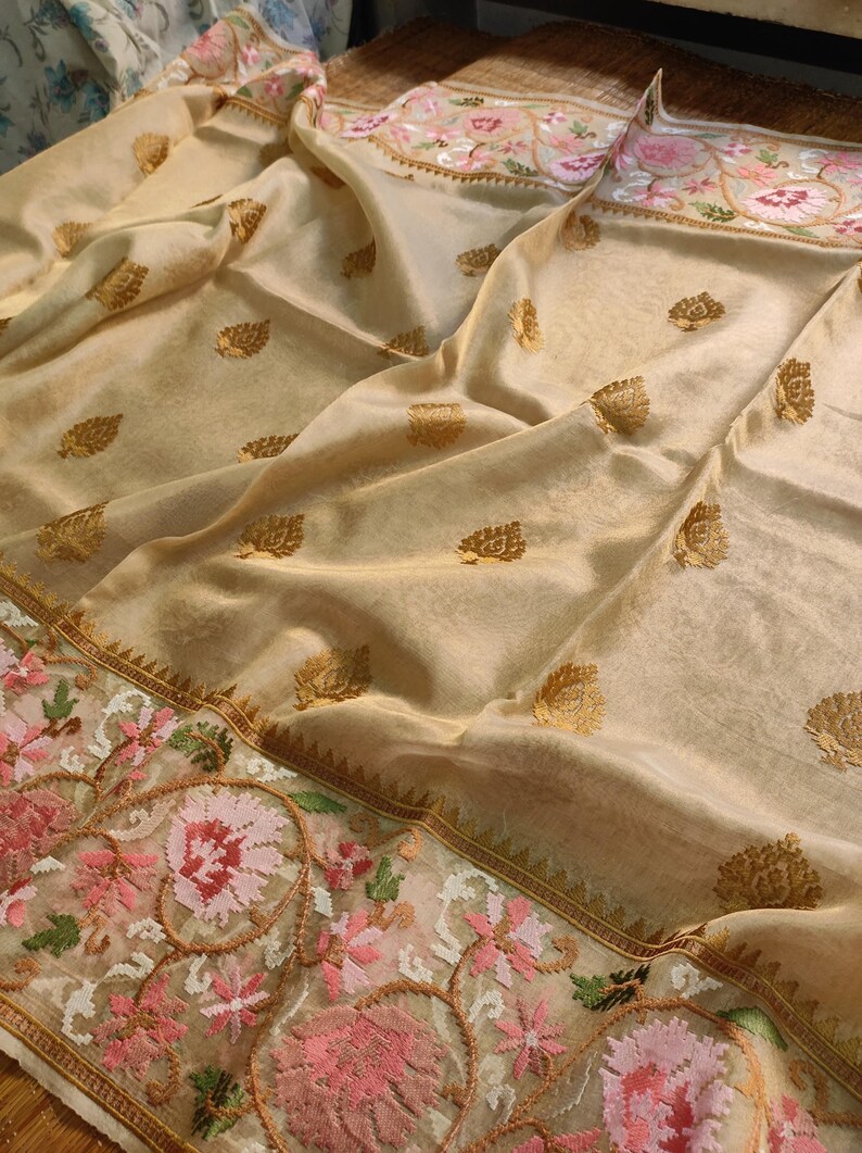 Pode incluir: Um sari bege com padr&otilde;es de folhas douradas e uma borda floral bordada em rosa, verde e branco. O tecido tem uma qualidade brilhante e transl&uacute;cida, com uma queda delicada.