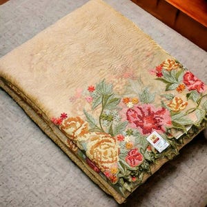 May include: Folded beige textile with intricate floral embroidery in shades of red, pink, orange, and green. The embroidered border features detailed flowers and leaves. The textile is folded neatly on a gray surface.
