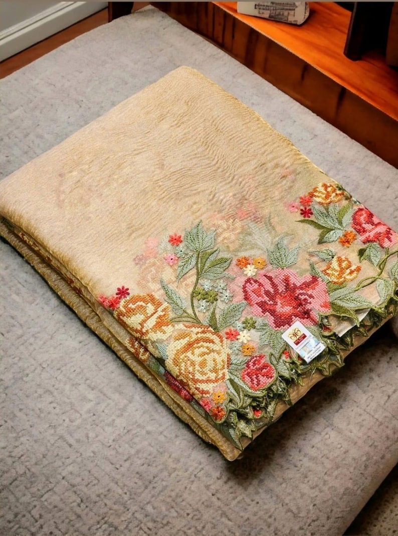 May include: Folded beige textile with intricate floral embroidery in shades of red, pink, orange, and green. The embroidered border features detailed flowers and leaves. The textile is folded neatly on a gray surface.