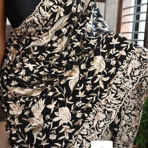 May include: Black textile with detailed beige floral and bird embroidery. The design features intricate patterns of flowers, leaves, and birds, creating a rich, textured appearance. The textile is draped, showcasing the detailed craftsmanship.