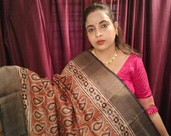 May include: A woman wearing a brown and black paisley patterned sari with a black border. The sari has a silver thread woven into the fabric. The woman is wearing a pink blouse.