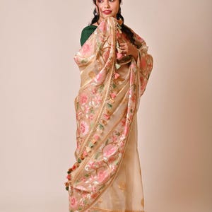 Pode incluir: Um sari bege com bordados florais intrincados em rosa e verde. O sari apresenta uma borda dourada e borlas decorativas. O tecido parece transparente e fluido, criando uma queda elegante. O sari &eacute; uma pe&ccedil;a de vestu&aacute;rio tradicional.