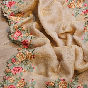 May include: A beige fabric with a textured surface and a floral embroidered border. The embroidery features pink, peach, and yellow roses with green leaves. The fabric is draped, showcasing its flowing quality.