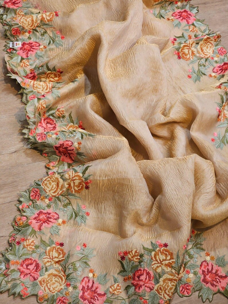 May include: A beige fabric with a textured surface and a floral embroidered border. The embroidery features pink, peach, and yellow roses with green leaves. The fabric is draped, showcasing its flowing quality.