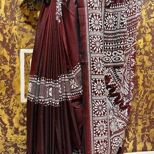 May include: A maroon saree with white floral and geometric patterns. The saree features detailed borders and a decorative hemline. The fabric appears to have a slight sheen, enhancing the rich color and design. The saree is draped, showcasing the intricate design.