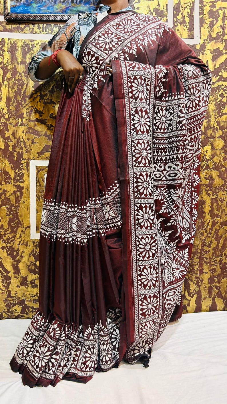May include: A maroon saree with white floral and geometric patterns. The saree features detailed borders and a decorative hemline. The fabric appears to have a slight sheen, enhancing the rich color and design. The saree is draped, showcasing the intricate design.