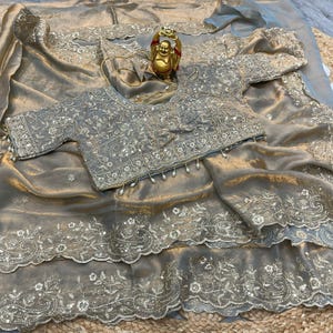 May include: A metallic gold saree featuring intricate silver embroidery and scalloped edges, paired with a matching short-sleeved blouse adorned with silver floral patterns, sequins, and small white beads hanging along the bottom hem.