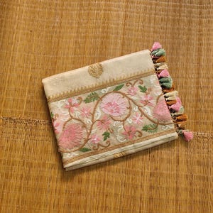 Pode incluir: Uma clutch bege com bordados florais intrincados em rosa, verde e castanho. A clutch apresenta um emblema dourado e &eacute; finalizada com uma borla de fios rosa, verdes e castanhos. Descansa sobre uma esteira tecida.