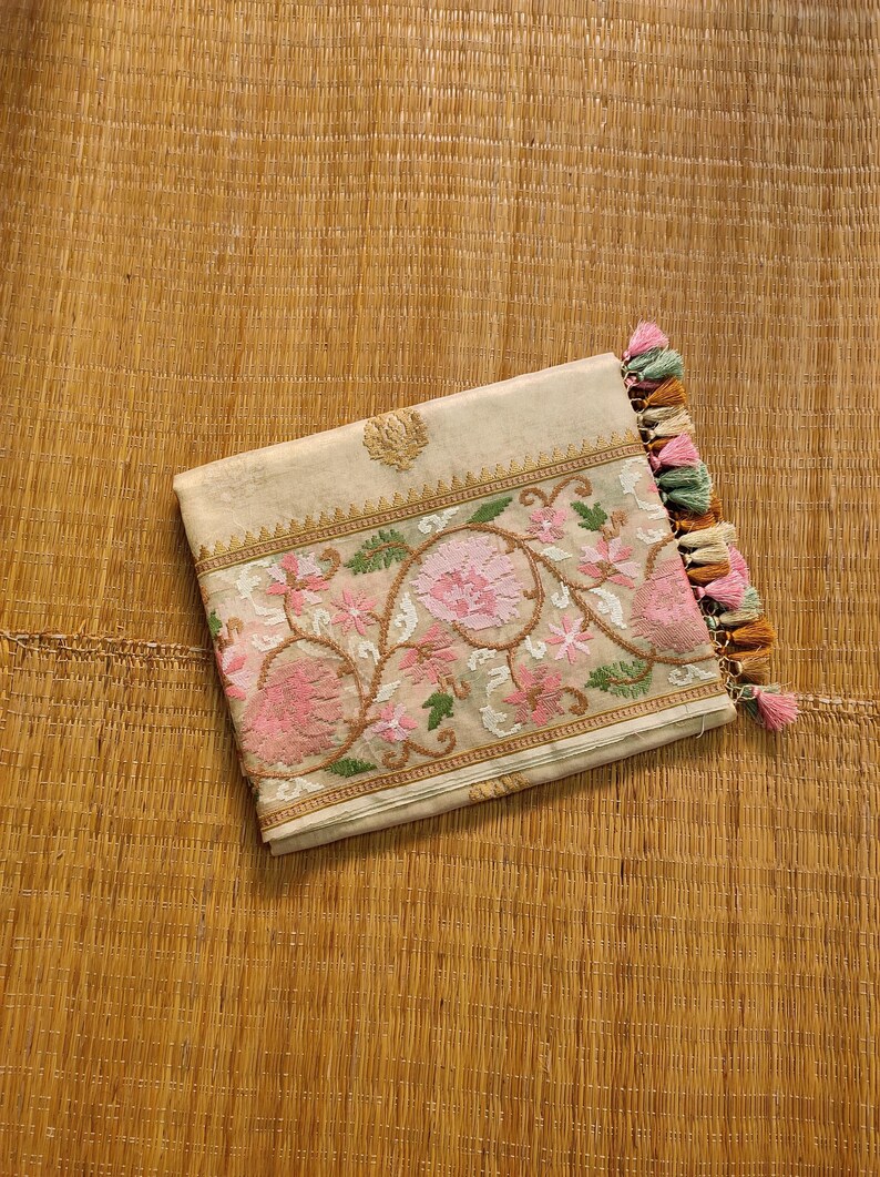 Pode incluir: Uma clutch bege com bordados florais intrincados em rosa, verde e castanho. A clutch apresenta um emblema dourado e &eacute; finalizada com uma borla de fios rosa, verdes e castanhos. Descansa sobre uma esteira tecida.