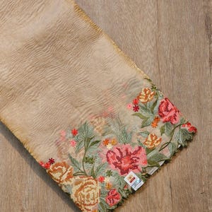 May include: A beige fabric with intricate floral embroidery. The embroidery features roses in shades of pink, orange, and red, with green leaves. The fabric has a sheer quality, and the embroidery is along the edge.