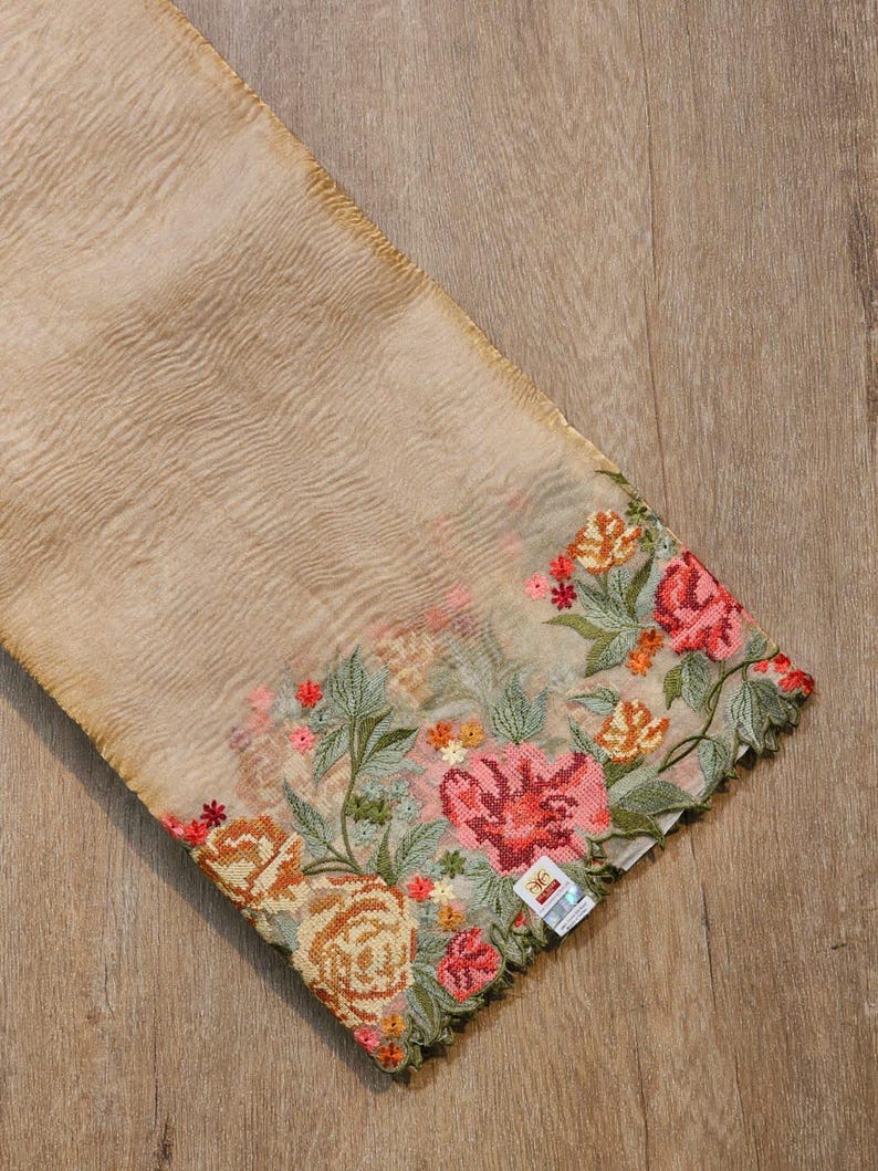 May include: A beige fabric with intricate floral embroidery. The embroidery features roses in shades of pink, orange, and red, with green leaves. The fabric has a sheer quality, and the embroidery is along the edge.