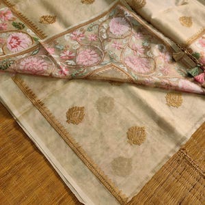 Pode incluir: T&ecirc;xtil bege com bordados florais intrincados em rosa e verde. O tecido apresenta detalhes dourados e uma borda detalhada. Uma borla com detalhes rosa e verde pende do canto. &Eacute; provavelmente um len&ccedil;o ou xale decorativo.