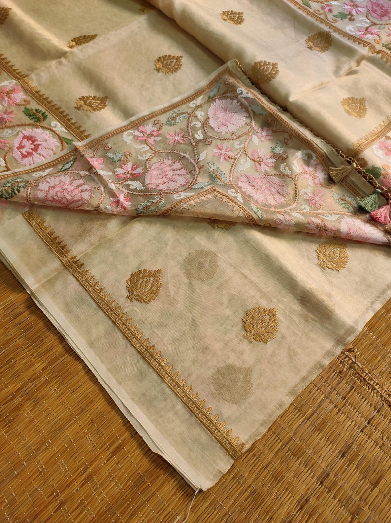 Pode incluir: T&ecirc;xtil bege com bordados florais intrincados em rosa e verde. O tecido apresenta detalhes dourados e uma borda detalhada. Uma borla com detalhes rosa e verde pende do canto. &Eacute; provavelmente um len&ccedil;o ou xale decorativo.