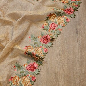 May include: A beige fabric with a textured surface, adorned with embroidered floral patterns. The floral design features roses in shades of pink, orange, and yellow, with green leaves. The edges of the fabric are scalloped.