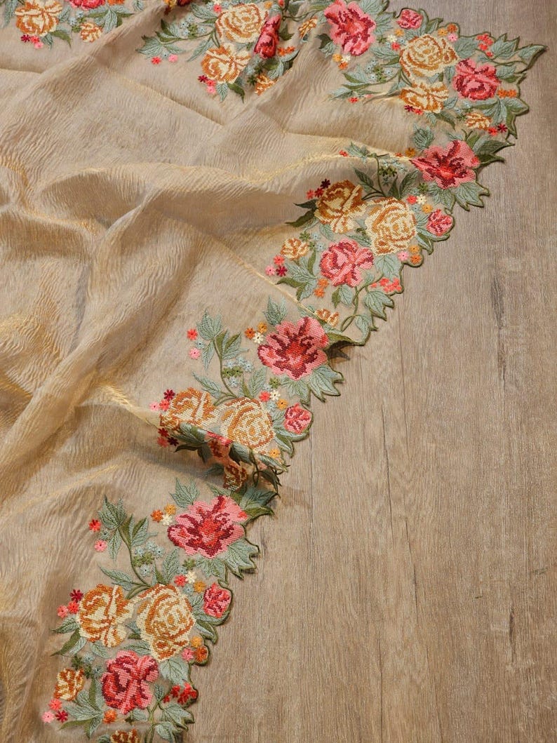 May include: A beige fabric with a textured surface, adorned with embroidered floral patterns. The floral design features roses in shades of pink, orange, and yellow, with green leaves. The edges of the fabric are scalloped.