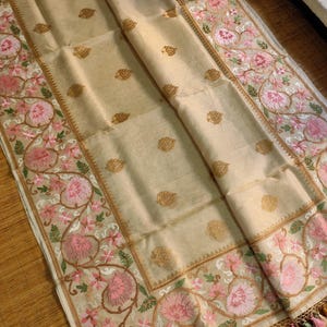 Pode incluir: T&ecirc;xtil bege com bordados intrincados. As bordas apresentam um desenho floral em rosa e verde, com detalhes dourados. O centro tem padr&otilde;es de folhas douradas. Tassels de v&aacute;rias cores adornam a borda inferior.