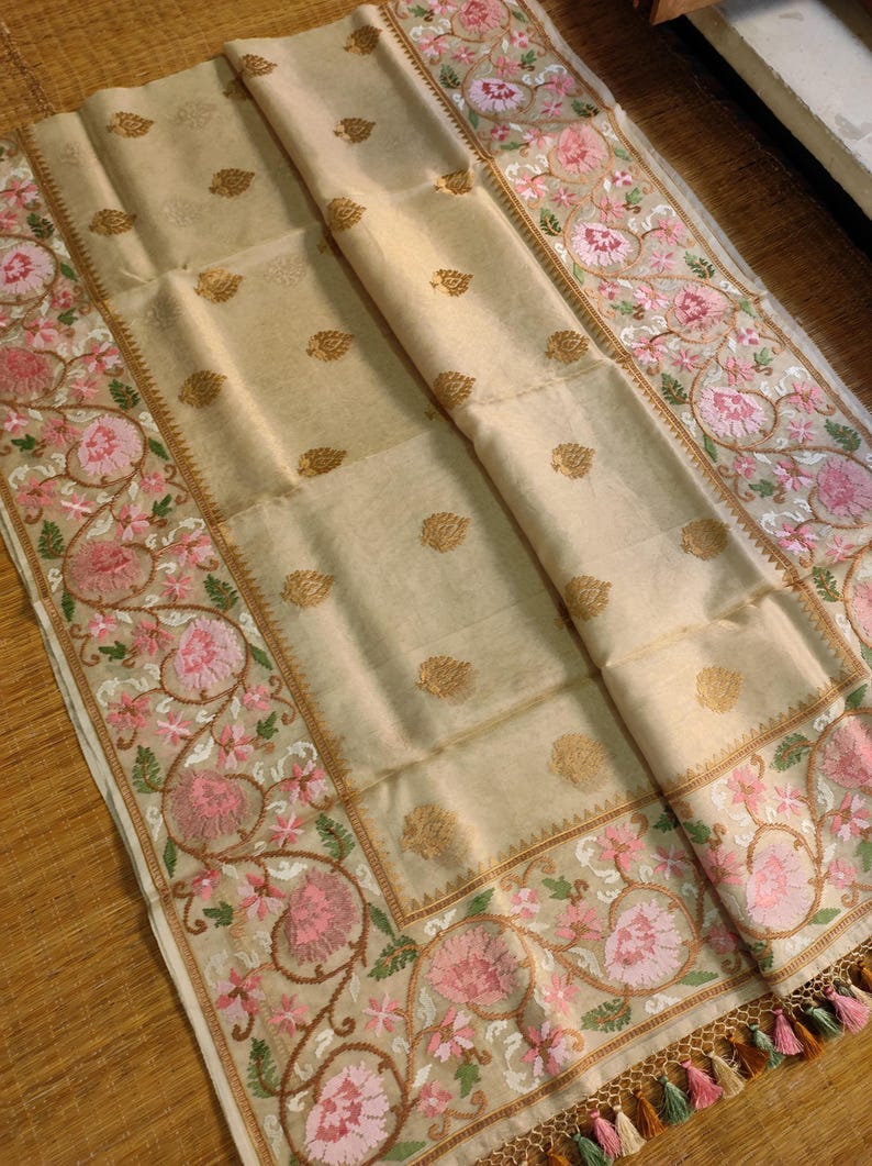 Pode incluir: T&ecirc;xtil bege com bordados intrincados. As bordas apresentam um desenho floral em rosa e verde, com detalhes dourados. O centro tem padr&otilde;es de folhas douradas. Tassels de v&aacute;rias cores adornam a borda inferior.