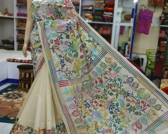 May include: Cream-colored sari with intricate floral embroidery in a variety of colors. The design features detailed stitching and a border with stripes. The sari is draped on a mannequin in a store setting.