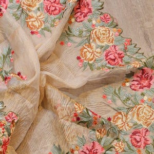 May include: A beige fabric with intricate floral embroidery in shades of pink, orange, and green. The embroidered flowers are arranged in a repeating pattern along the edge of the fabric, creating a decorative border. The fabric has a delicate, flowing texture.