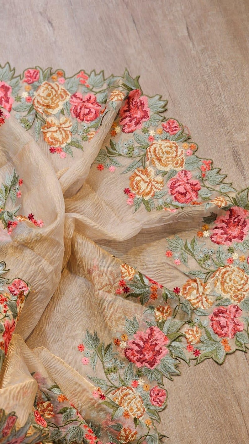 May include: A beige fabric with intricate floral embroidery in shades of pink, orange, and green. The embroidered flowers are arranged in a repeating pattern along the edge of the fabric, creating a decorative border. The fabric has a delicate, flowing texture.
