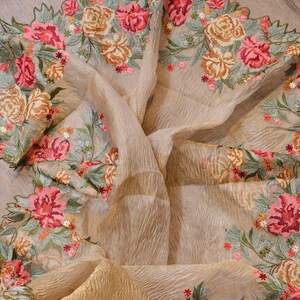 May include: A beige, sheer fabric with intricate floral embroidery. The embroidery features pink, orange, and red roses with green leaves. The fabric is draped, showcasing its texture and the detailed floral design.
