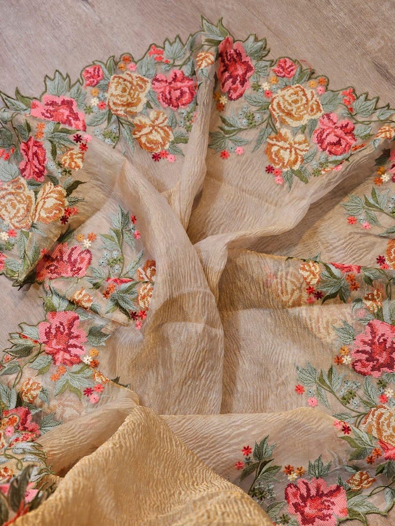 May include: A beige, sheer fabric with intricate floral embroidery. The embroidery features pink, orange, and red roses with green leaves. The fabric is draped, showcasing its texture and the detailed floral design.