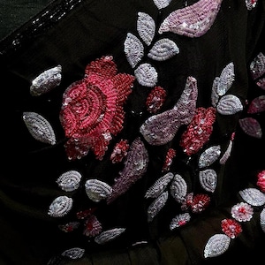 May include: A black saree featuring intricate embroidery with red and silver sequins. The fabric is adorned with floral and leaf motifs created from dense beadwork and metallic thread, complemented by a black sequined border along the edge.