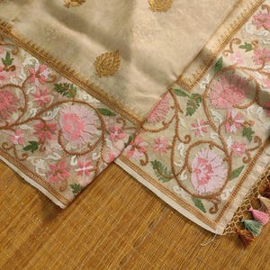 Pode incluir: T&ecirc;xtil creme com padr&otilde;es de folhas douradas e bordados florais em rosa, verde e castanho. A borda apresenta intrincados desenhos florais e uma borda dourada. Tassels em v&aacute;rias cores s&atilde;o vis&iacute;veis.